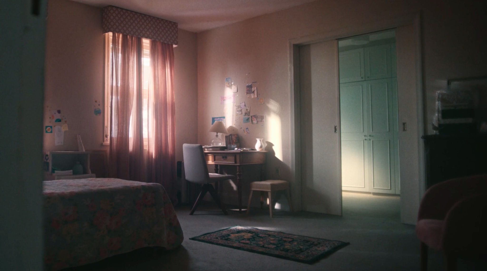 Quarto com paredes rosadas, cortina, escrivaninha e porta verde-clara.