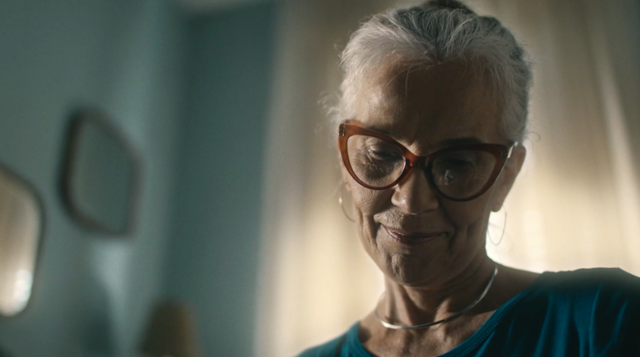 Mulher de óculos e blusa azul sorrindo, em um quarto com paredes verdes.