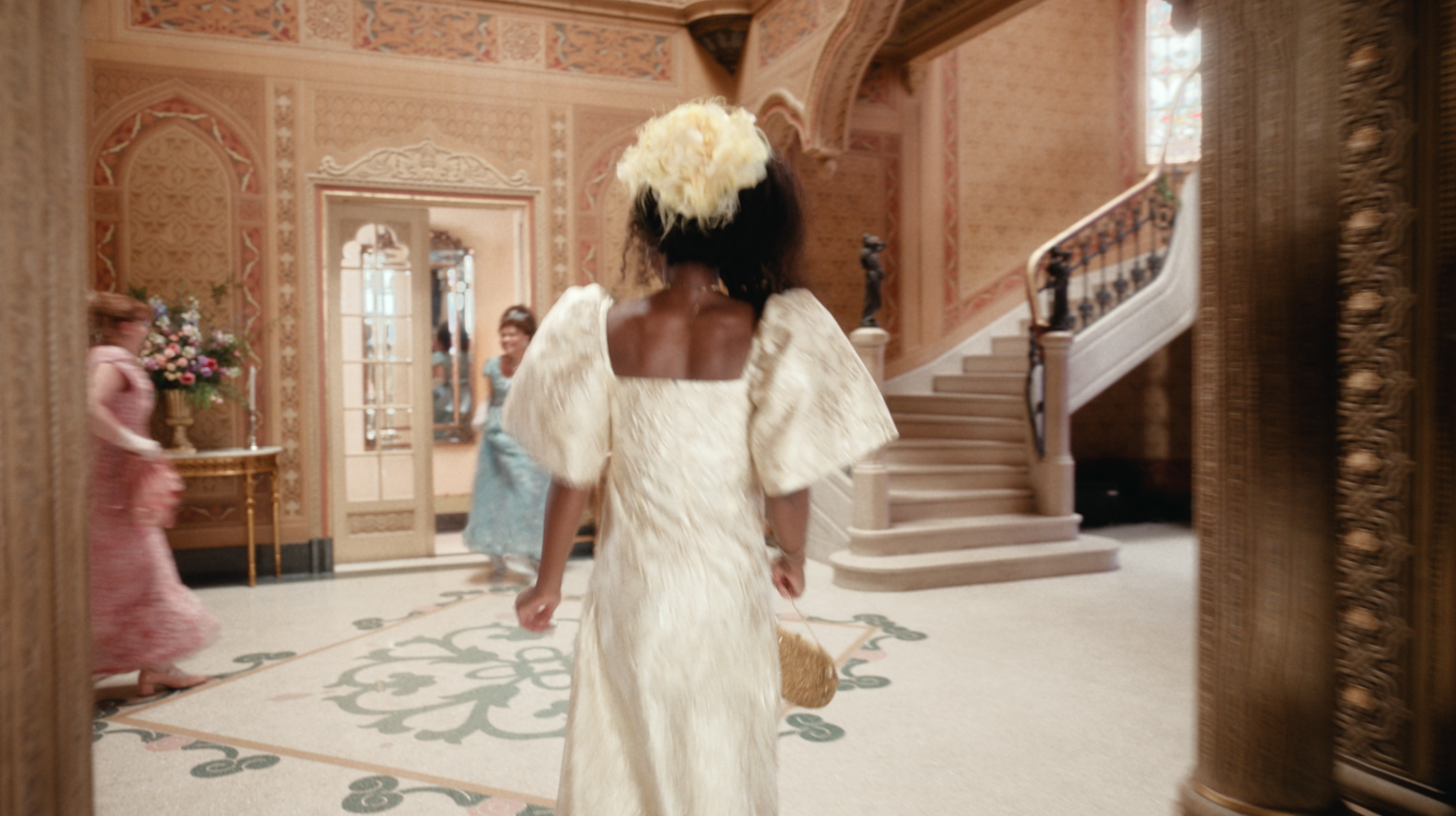 Mulher de vestido claro em um hall com escadaria e paredes ornamentadas.