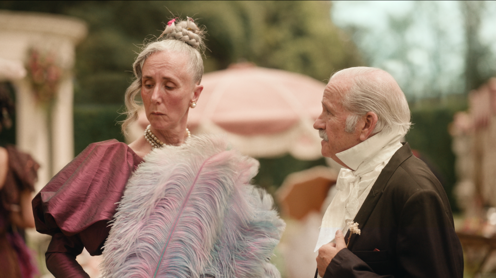 Mulher com leque de plumas ao lado de um homem em traje de época no jardim.
