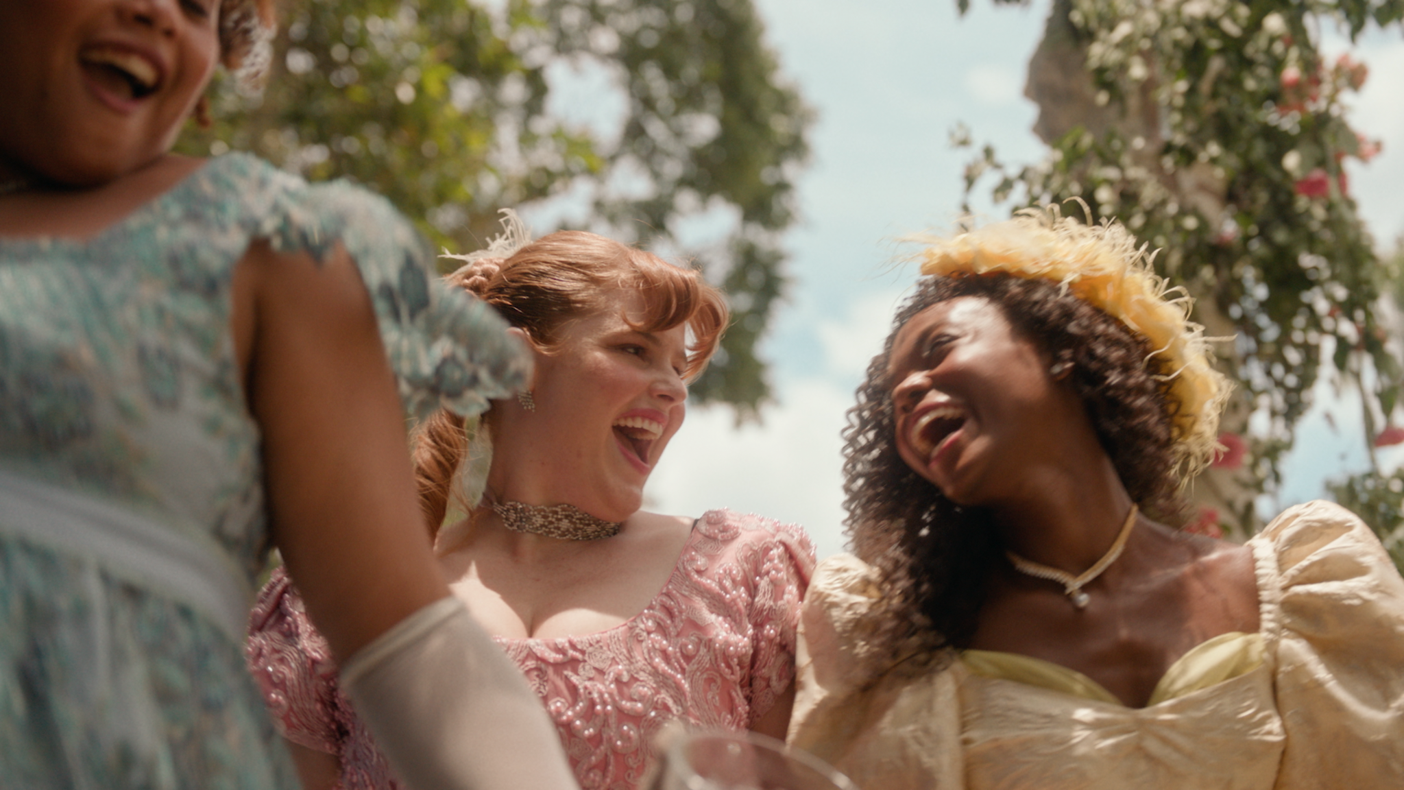 Mulheres com vestidos de época e flores nos cabelos sorrindo no jardim.