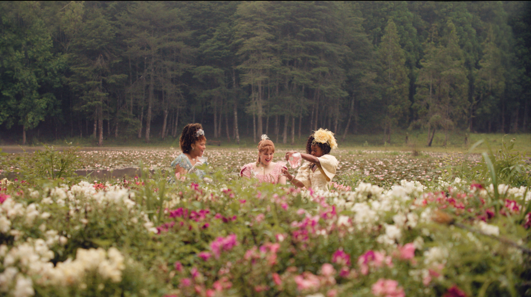Mulheres entre flores em um campo cercado por uma floresta.