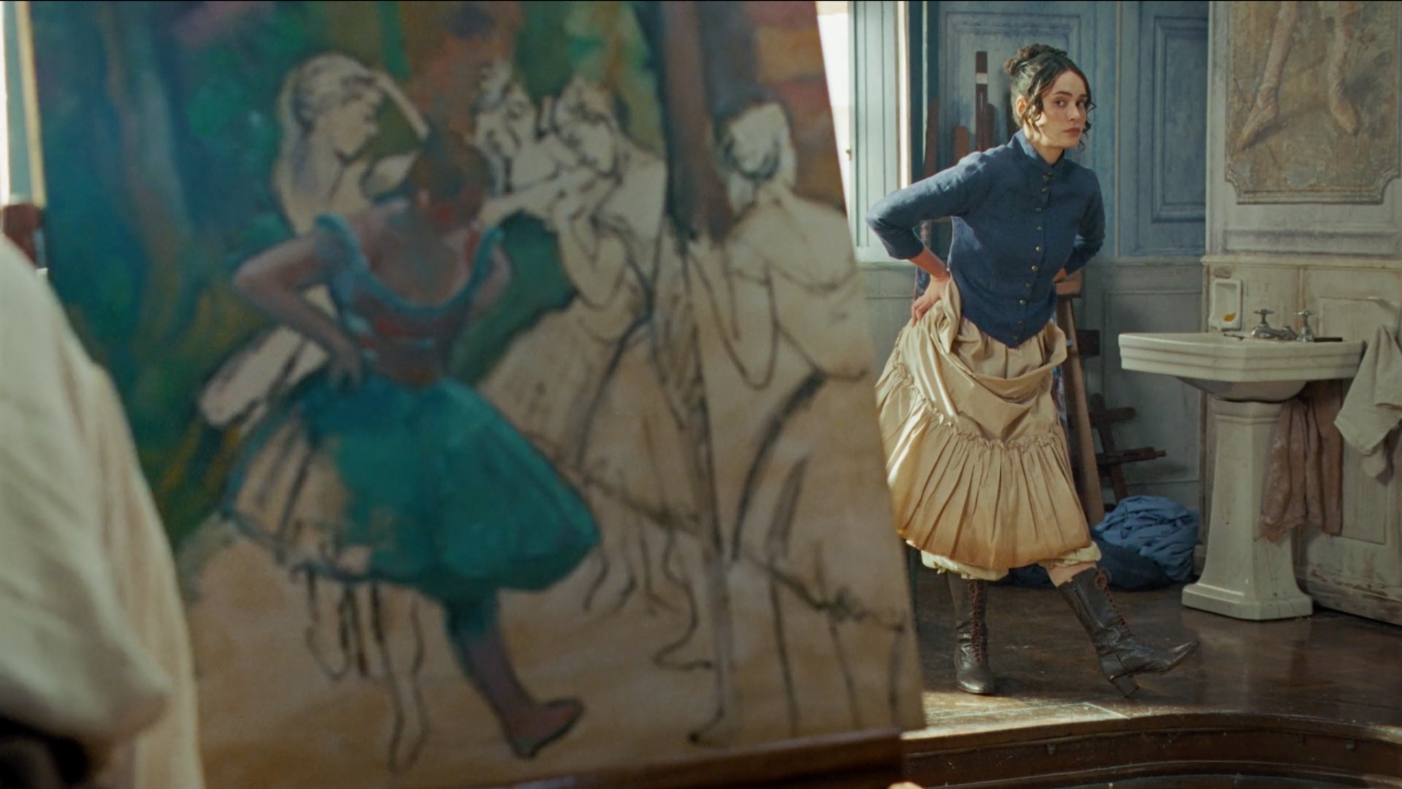 Mulher com blusa azul e saia clara ao lado de uma pintura de bailarinas, em frente a uma pia.