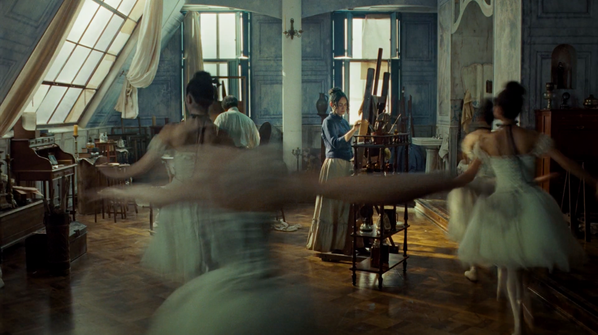 Bailarinas dançando entre os cavaletes de um ateliê com grandes janelas.