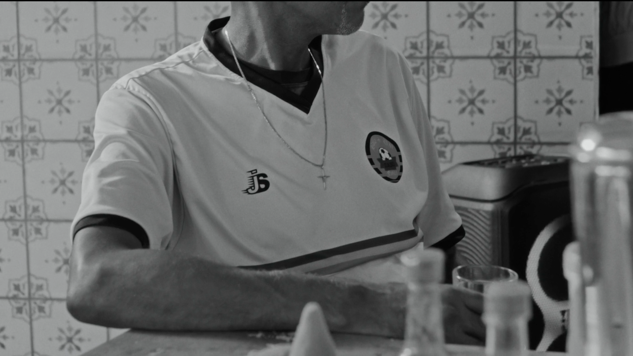 Detalhe de um homem com camisa de futebol e de um copo sobre a mesa do bar, em preto e branco.