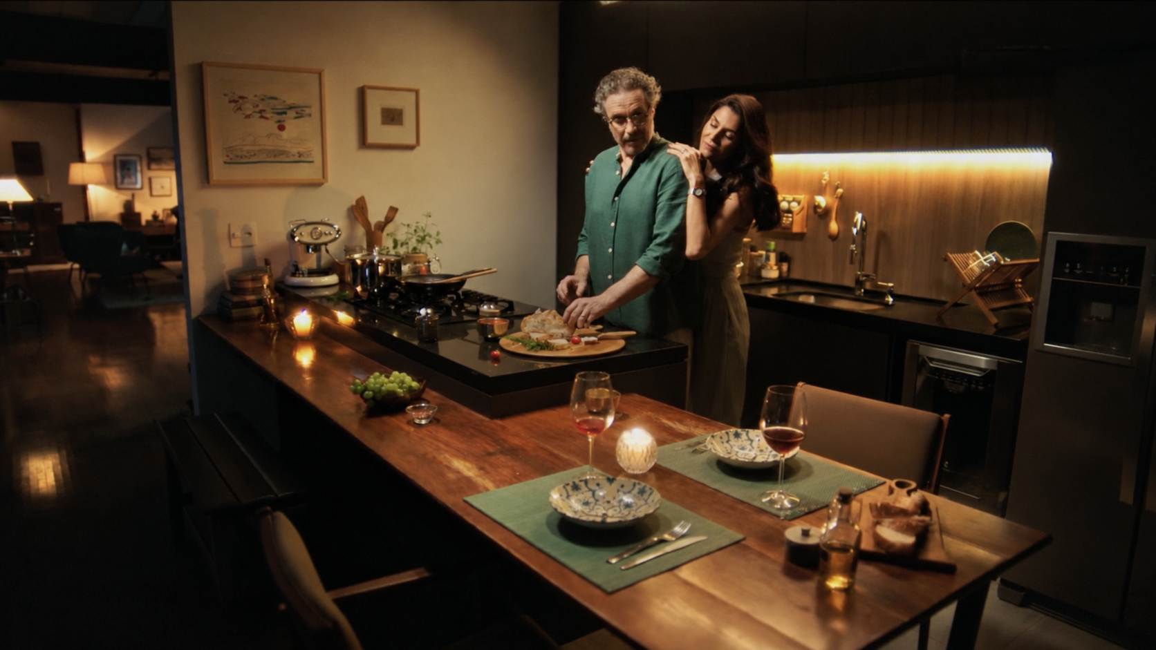 Casal junto à ilha da cozinha sob luz quente, com pratos, taças e velas em primeiro plano.