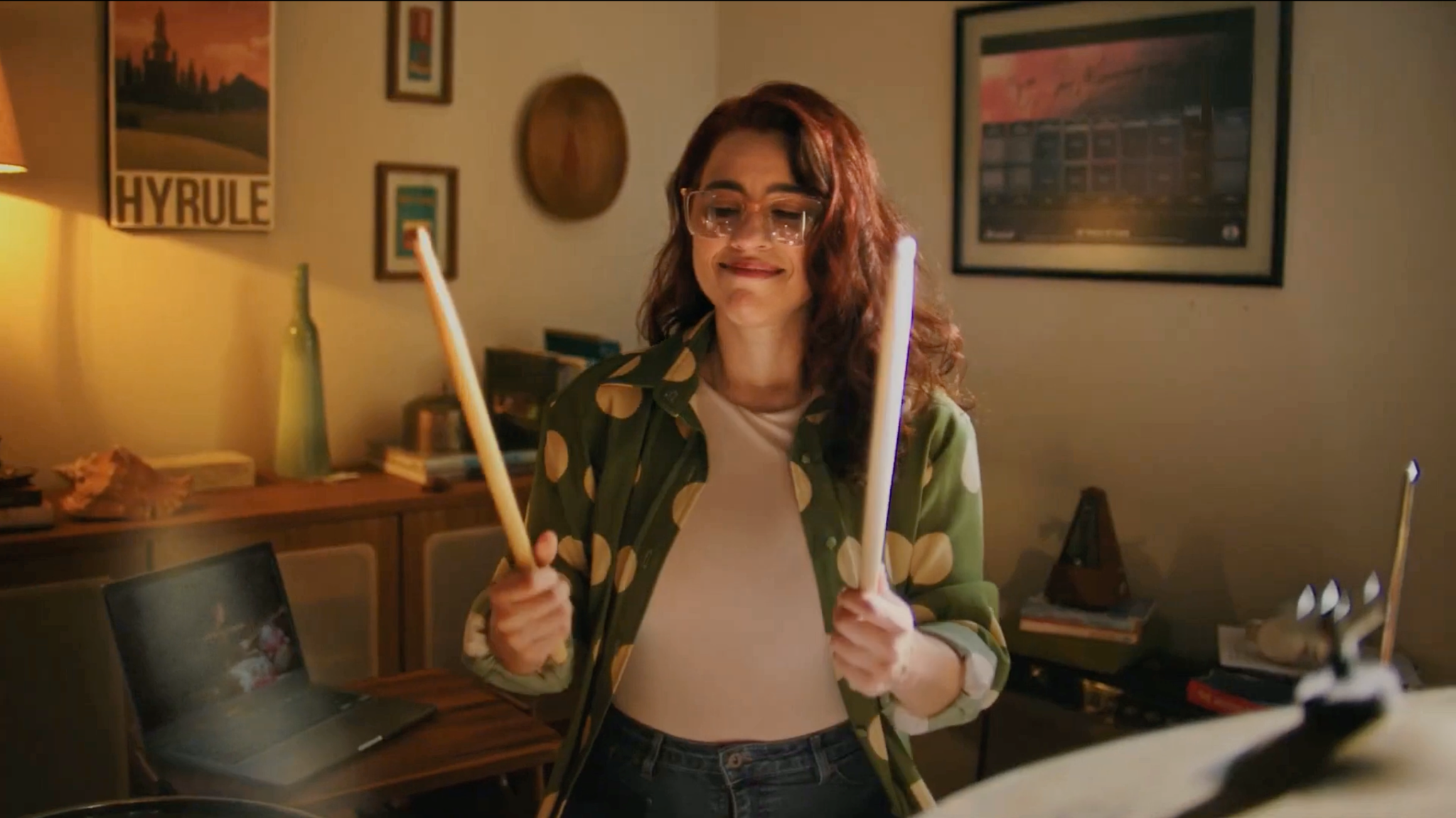Baterista de óculos segurando baquetas em um quarto com quadros e pôster na parede.