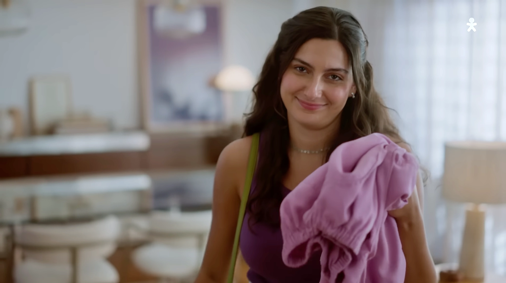 Mulher com uma blusa roxa, segurando um casaco lilás em uma sala clara.